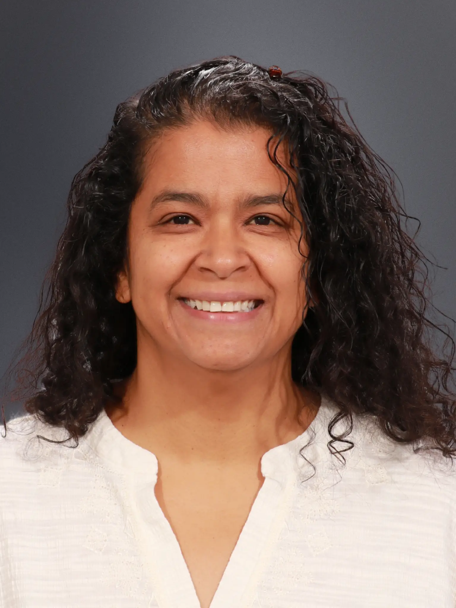 Smiling woman with dark curly hair wearing a white blouse against a gray backdrop.