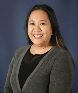 Smiling woman in a gray cardigan and black top, posing against a blue background.